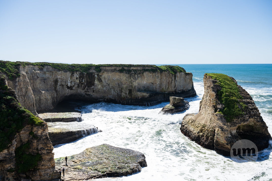 Dramatic California coastline featuring rugged cliffs plunging into the turbulent Pacific Ocean, with a few small figures standing on a rocky outcrop observing the crashing waves under a clear blue sky.