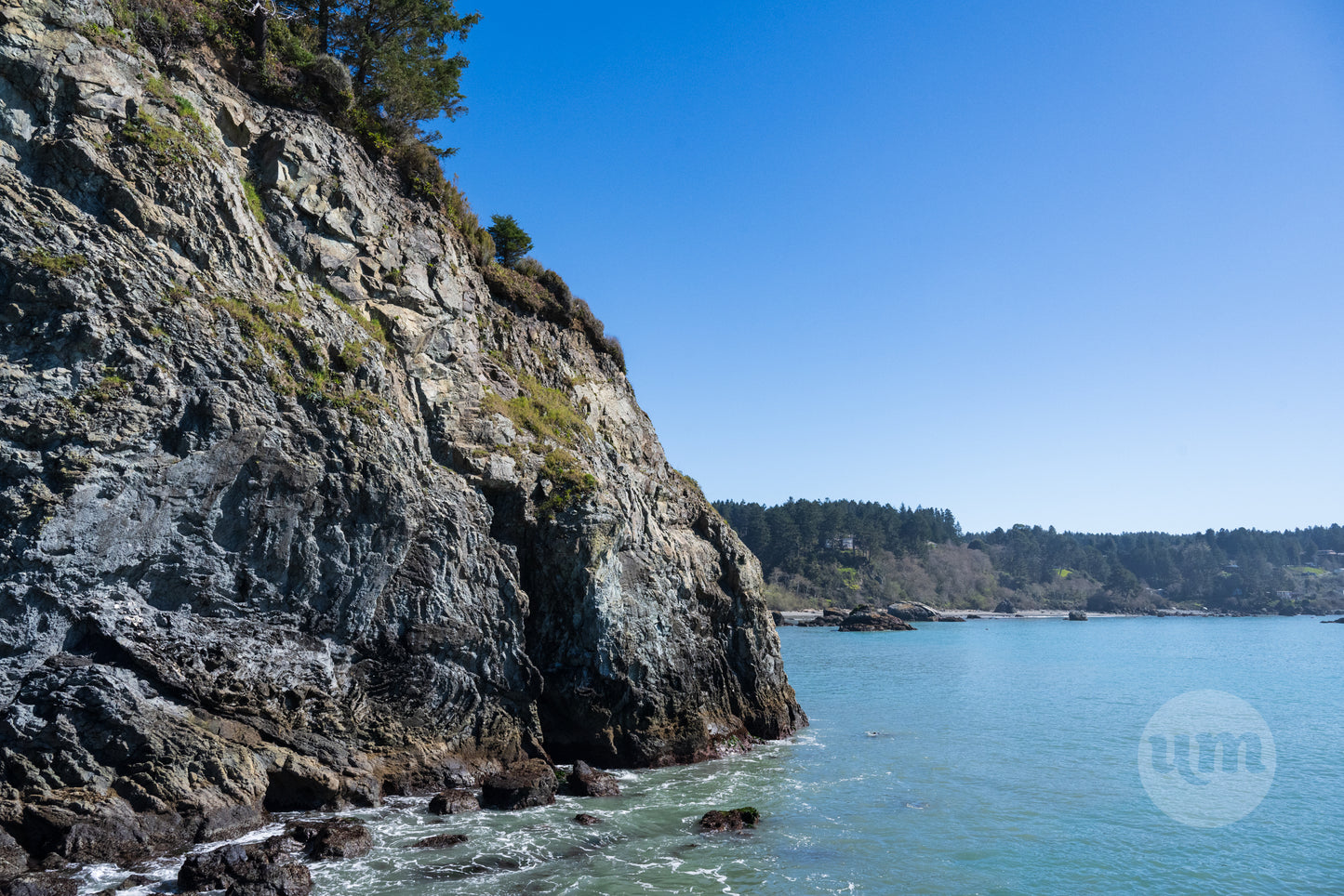 A photograph depicting a rugged Oregon Coast headland with cliffs plunging into the clear blue Pacific Ocean, with a bright, sunny sky above and forested coastline in the distance.