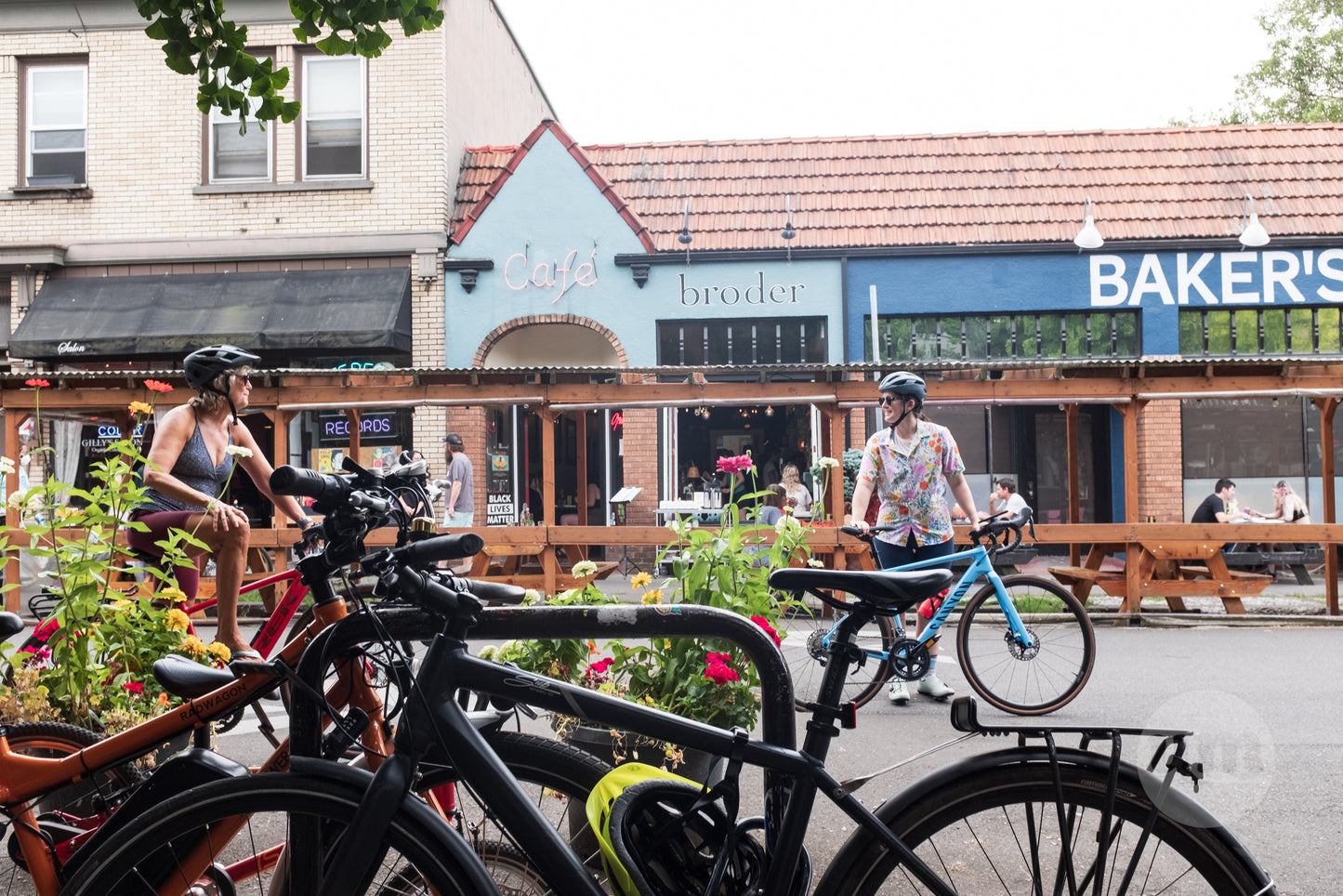 A vibrant summer street scene in Portland, Oregon, featuring cyclists, pedestrians, and local businesses with outdoor seating and colorful awnings.