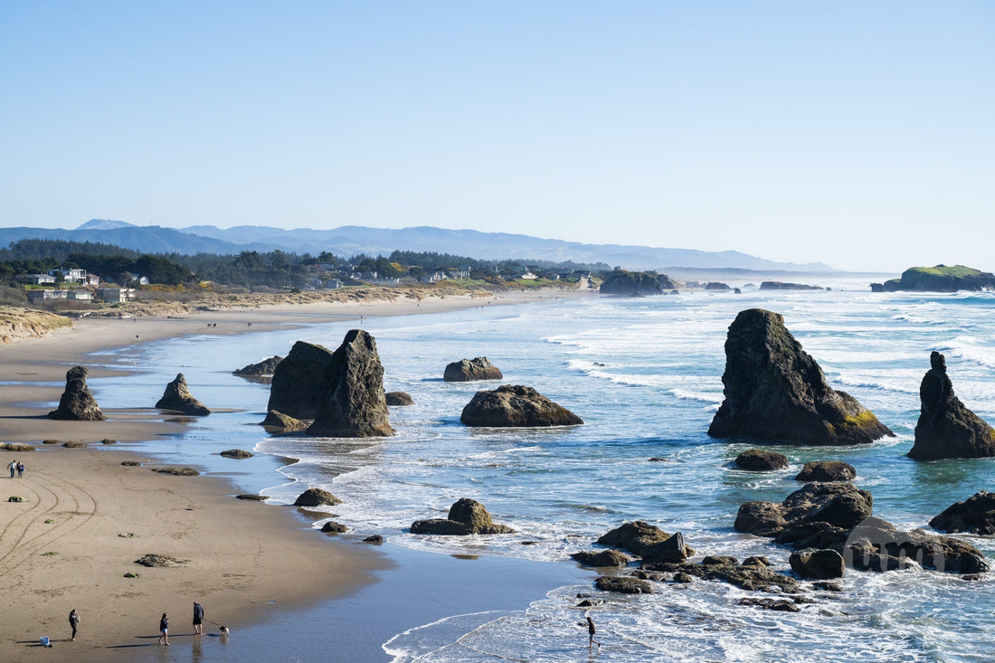Oregon Coast scenic vista featuring dramatic sea stacks rising from the Pacific Ocean, sandy beach with distant figures, and hazy coastal mountains under a clear blue sky.