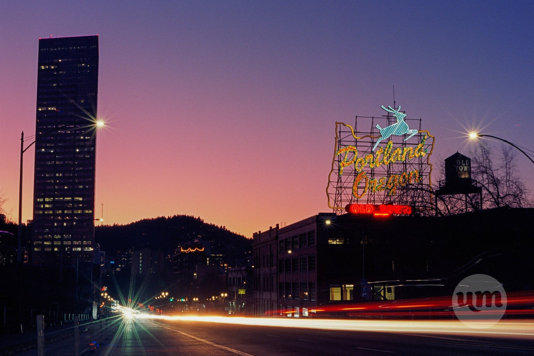 A vibrant Portland, Oregon cityscape at twilight, featuring the iconic "Portland Oregon" sign with its glowing stag, a tall illuminated skyscraper, and dramatic light trails from vehicles on the street, all set against a colorful purple and orange sky.
