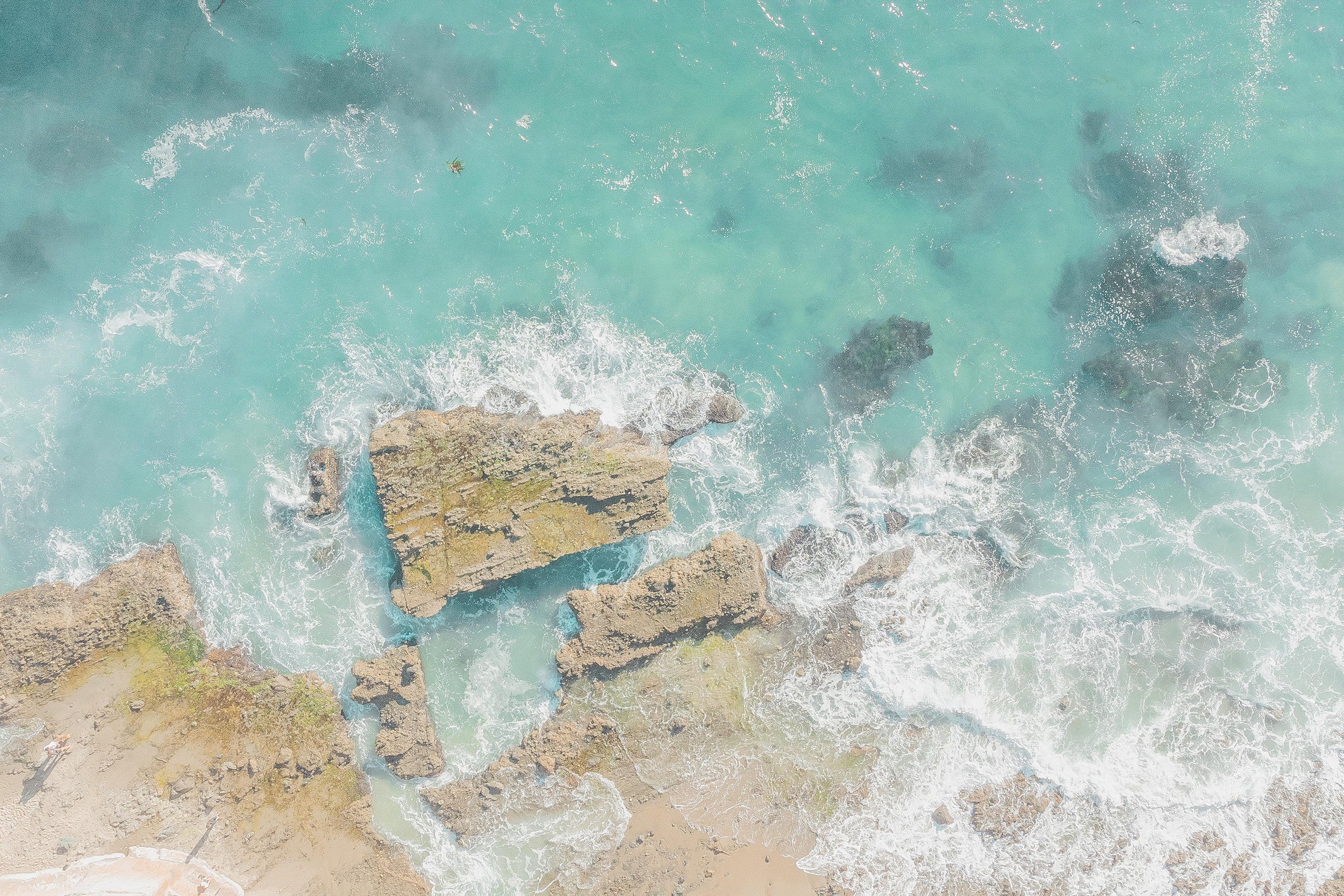Aerial view of ocean waves crashing against rocks with clear blue water.