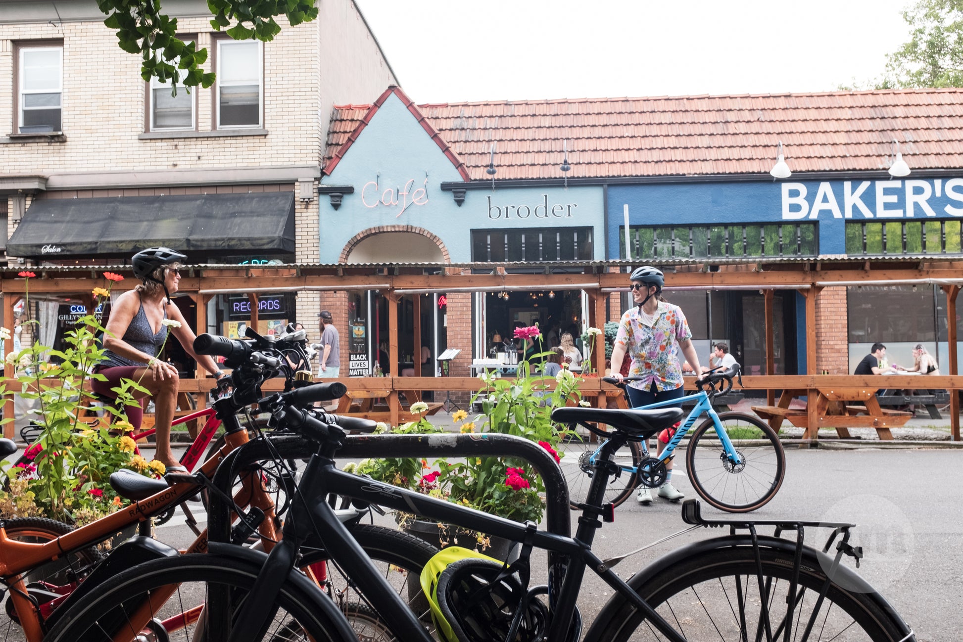 A vibrant summer street scene in Portland, Oregon, featuring cyclists, pedestrians, and local businesses with outdoor seating and colorful awnings.